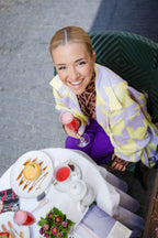 A woman in a patterned jacket holds a drink at a table with tea, a dessert, and flowers.