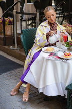 Woman wearing a yellow and lavender coat, leopard print turtleneck, and purple pants, pouring tea at an outdoor cafe.