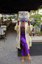 A person in a leopard print turtleneck, a yellow and lavender coat, and purple pants reading a newspaper while sitting on a bench.