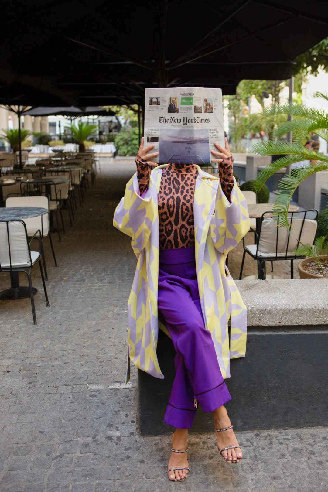 A person in a leopard print turtleneck, a yellow and lavender coat, and purple pants reading a newspaper while sitting on a bench.