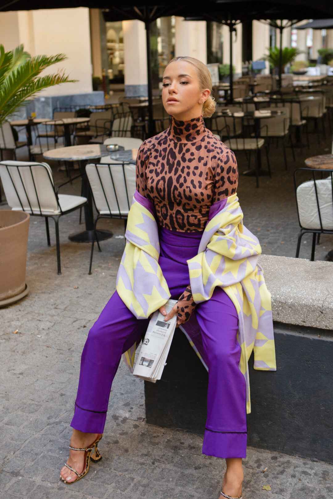 A woman in a leopard-print turtleneck and purple pants, holding a newspaper and sitting outdoors.
