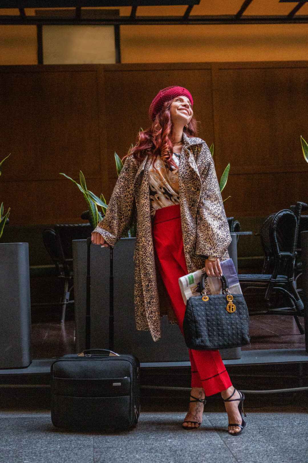 A stylish woman in a leopard-print coat, red pants, beret, and sandals stands next to her suitcase in a public space. She holds a black purse and a newspaper.