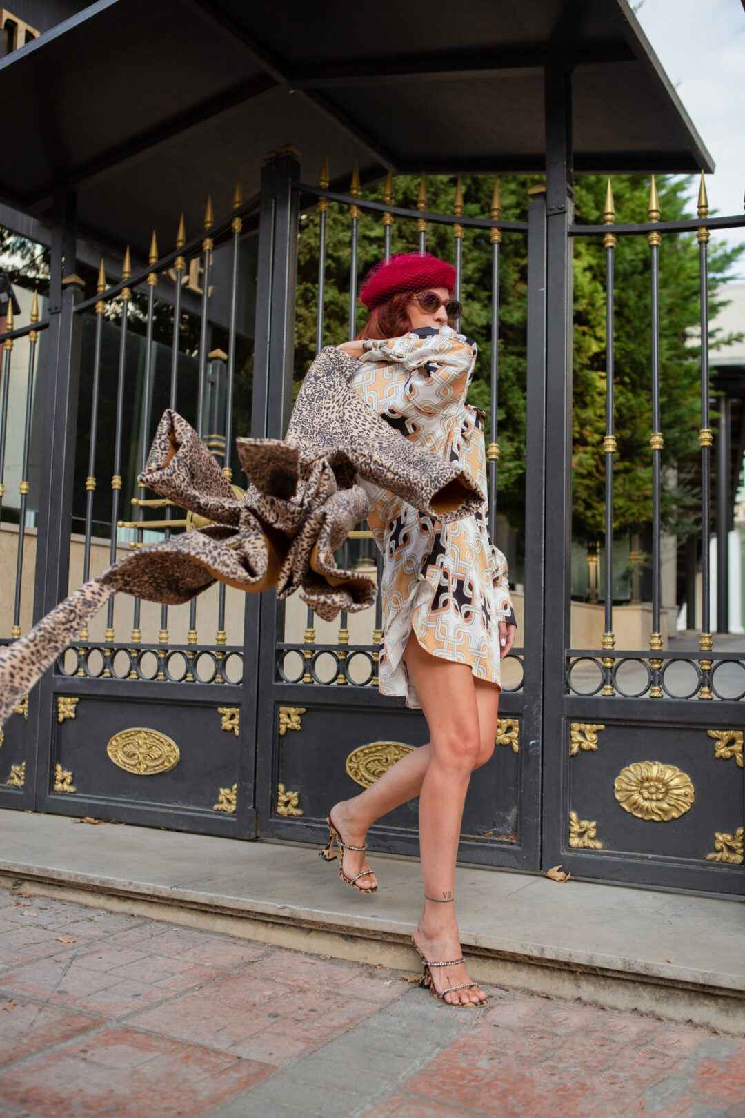 Woman in patterned shirt dress, beret, and high heels walking in front of a black and gold fence.