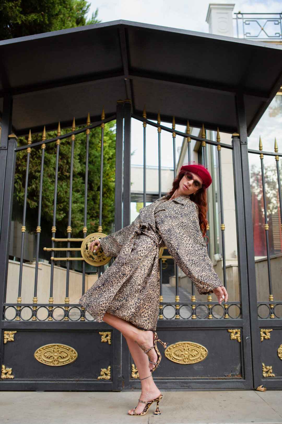 A woman poses in a leopard print coat and matching heels, wearing a red beret and sunglasses in front of a black and gold gate.
