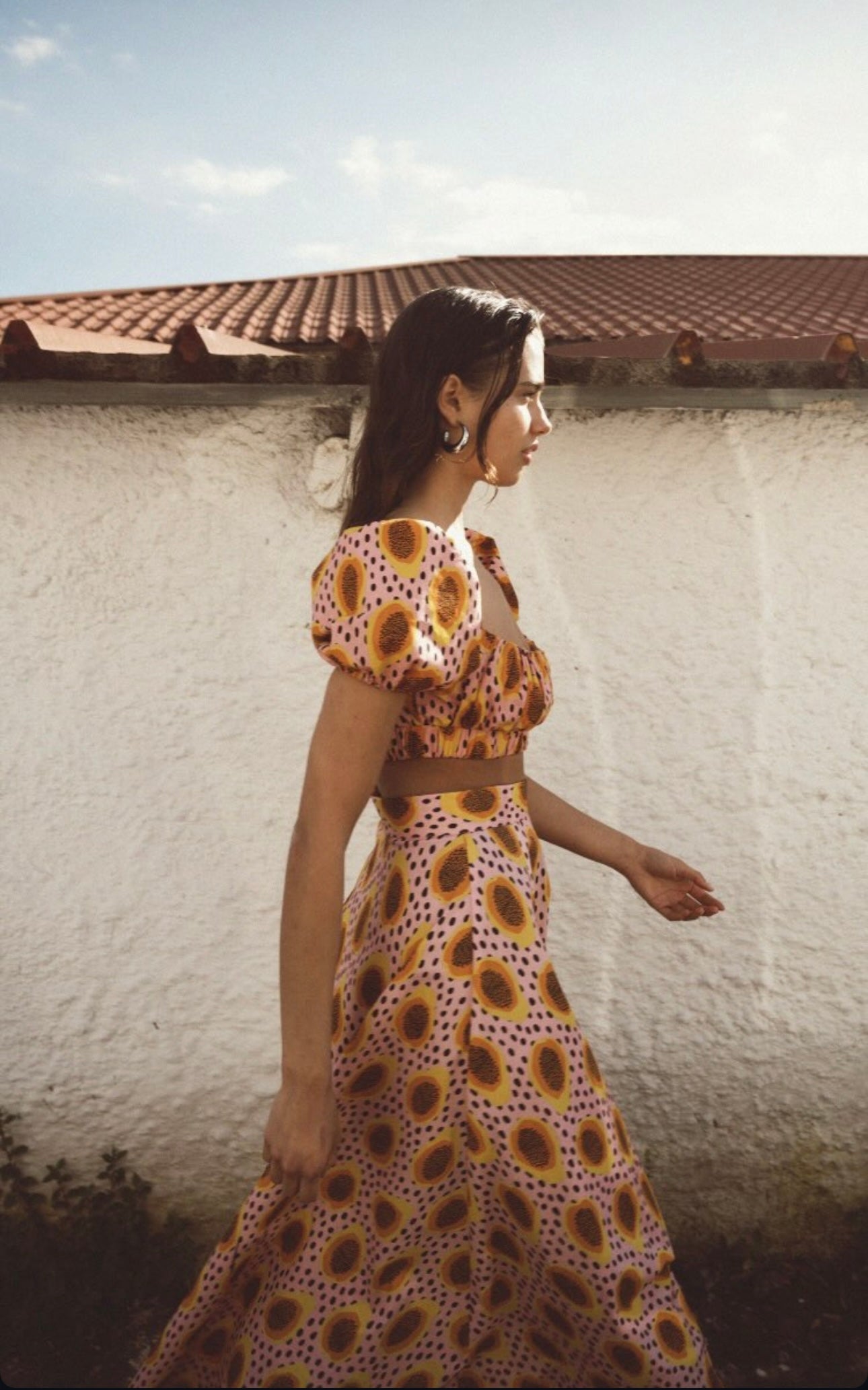 Woman wearing a patterned dress standing against a white wall with a red-tiled roof.