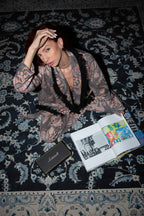 Person with tattoos lying on a patterned rug with an open book and smartphone.