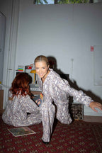 Two women in snakeskin print jumpsuits pose in a room with records and vintage electronics on a red patterned rug.