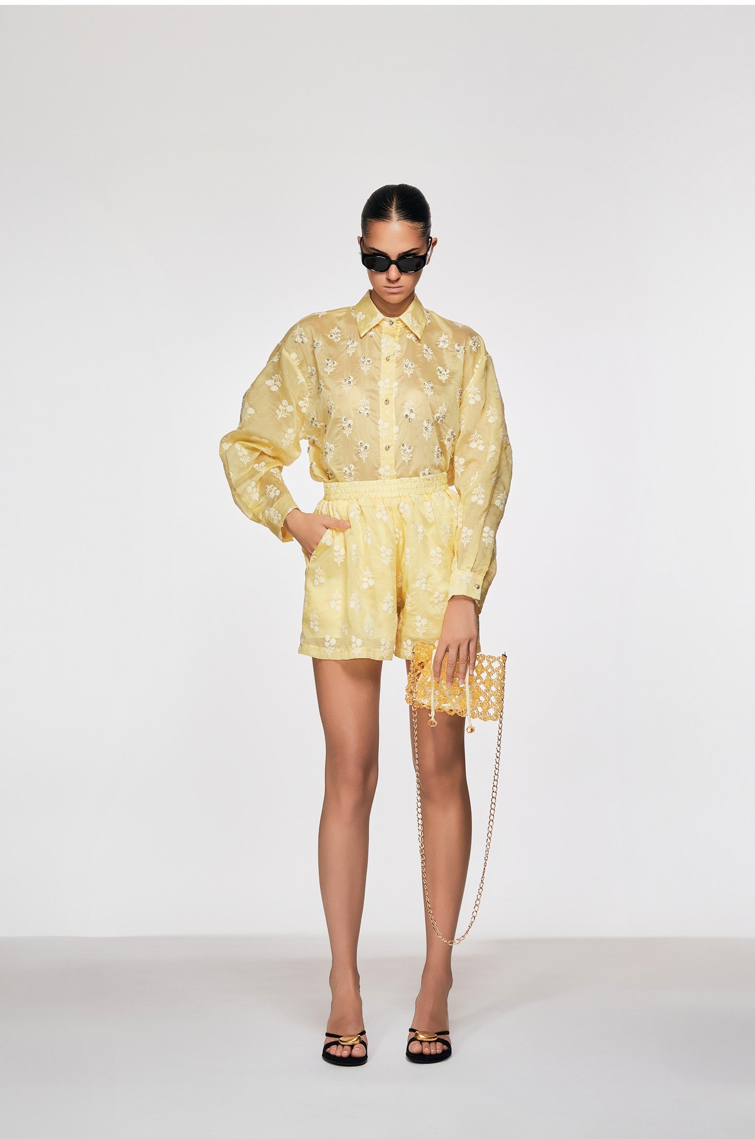 Person wearing a yellow outfit with sunglasses and holding a clutch against a white background