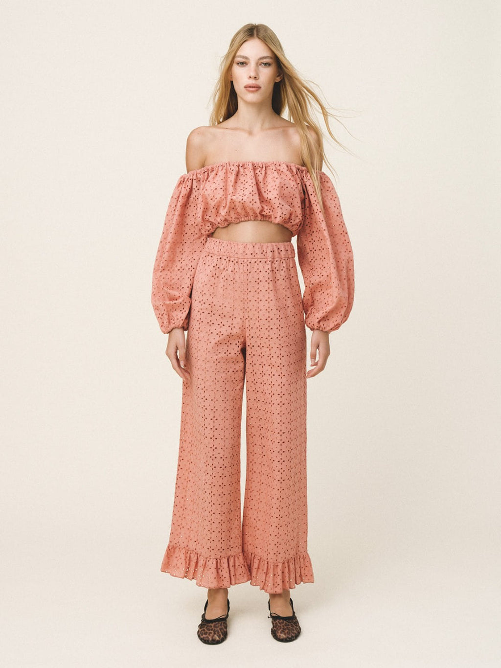 A woman modeling a matching two-piece outfit consisting of an off-the-shoulder cropped top with long puff sleeves and high-waisted wide-leg pants, both in a textured dusty rose pink fabric, set against a plain light beige studio background.
