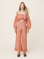 A woman modeling a matching two-piece outfit consisting of an off-the-shoulder cropped top with long puff sleeves and high-waisted wide-leg pants, both in a textured dusty rose pink fabric, set against a plain light beige studio background.