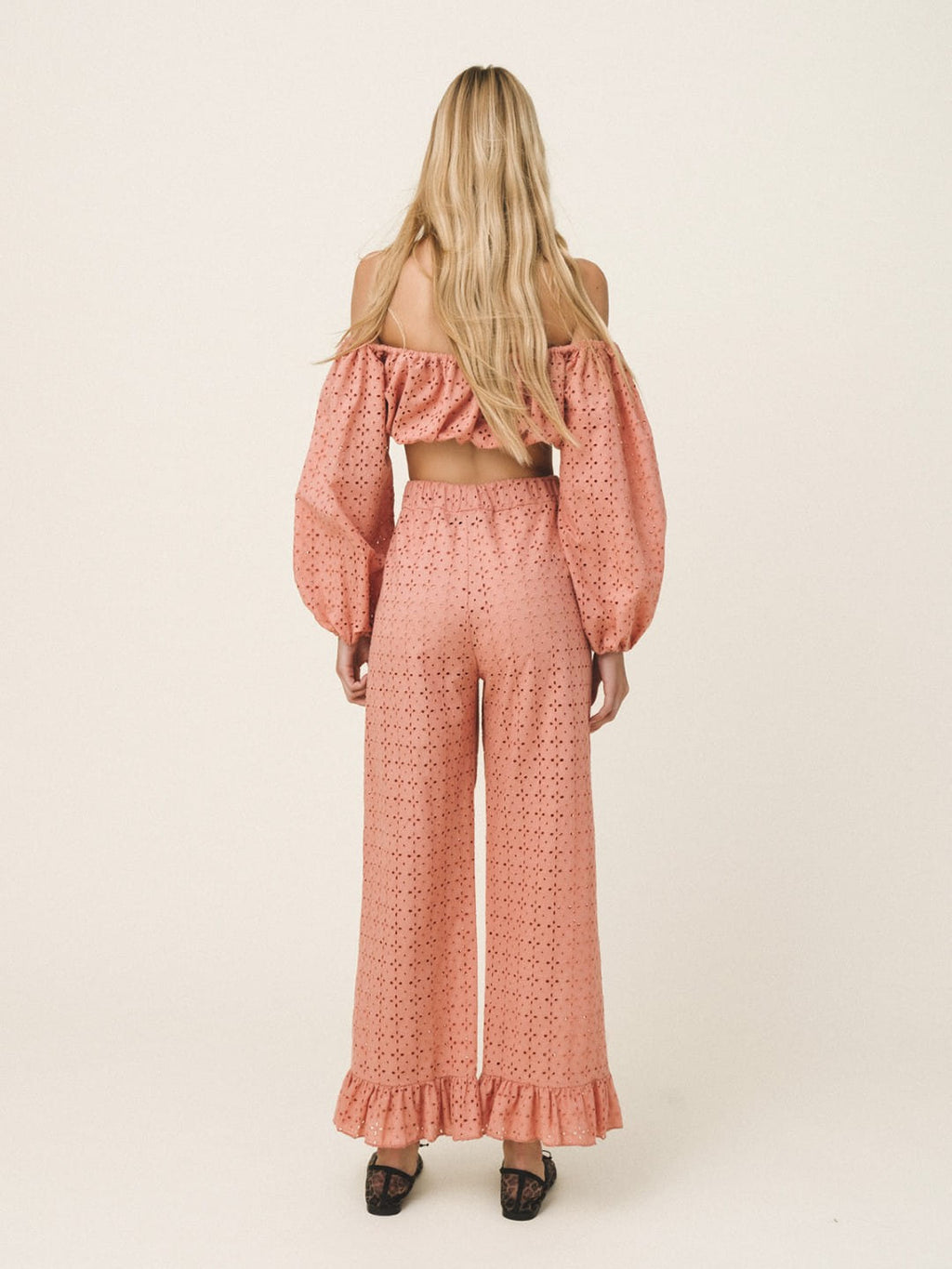 A woman viewed from behind wearing a matching two-piece set consisting of an off-the-shoulder long-sleeve crop top and high-waisted wide-leg pants, both in a dusty rose pink eyelet fabric. She is wearing black pointed-toe heels against a plain, warm off-white studio background.
