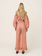 A woman viewed from behind wearing a matching two-piece set consisting of an off-the-shoulder long-sleeve crop top and high-waisted wide-leg pants, both in a dusty rose pink eyelet fabric. She is wearing black pointed-toe heels against a plain, warm off-white studio background.
