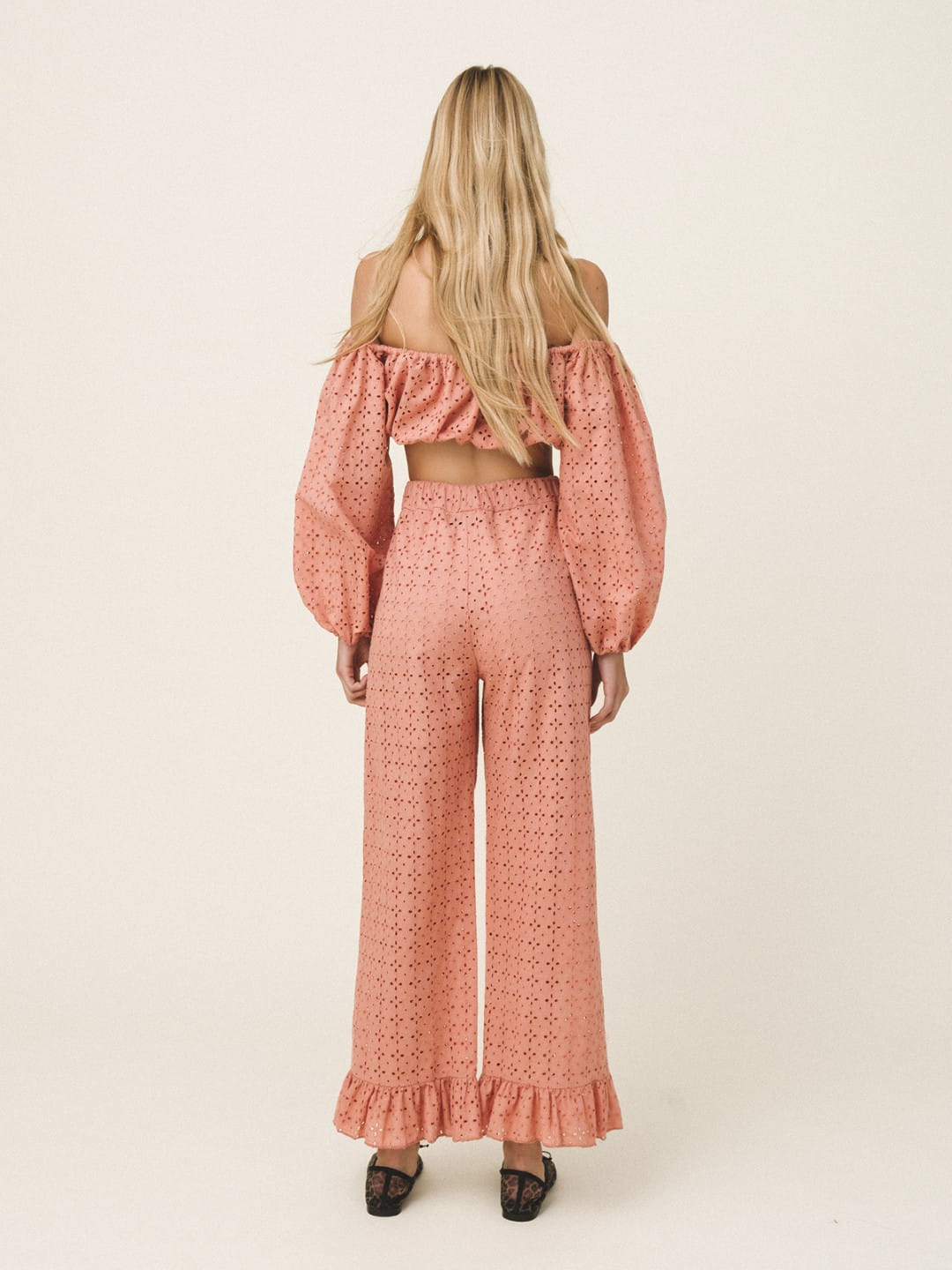 A woman viewed from behind wearing a matching two-piece set consisting of an off-the-shoulder long-sleeve crop top and high-waisted wide-leg pants, both in a dusty rose pink eyelet fabric. She is wearing black pointed-toe heels against a plain, warm off-white studio background.