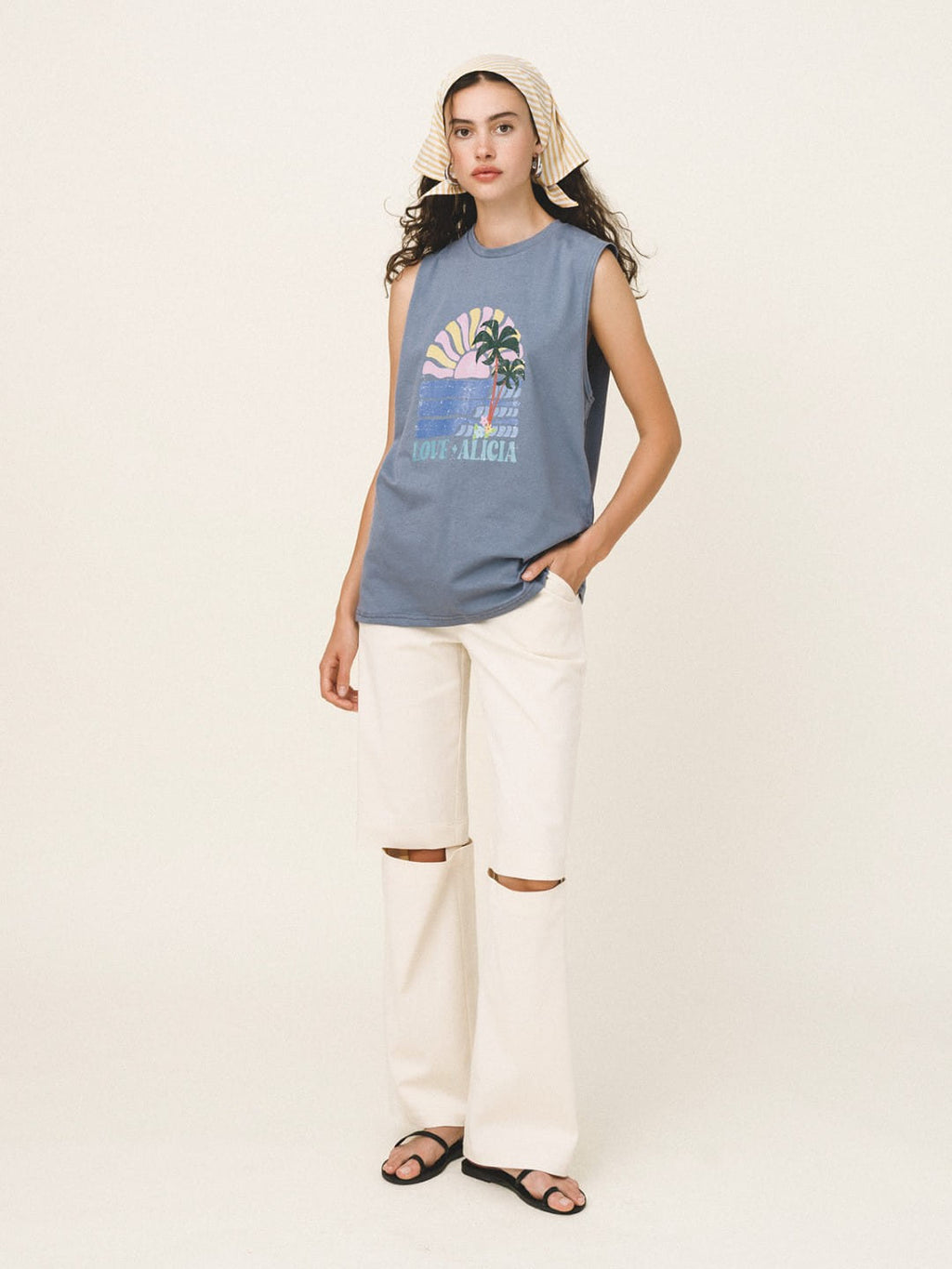 Woman wearing a sleeveless graphic shirt and white pants on a plain background