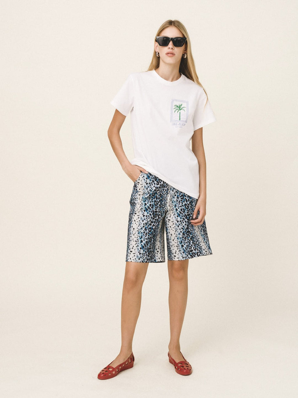 A woman modeling a white short-sleeve t-shirt with a small palm tree graphic on the chest, paired with blue and white patterned shorts and red flat shoes, against a solid light beige background.