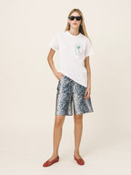 A woman modeling a white short-sleeve t-shirt with a small palm tree graphic on the chest, paired with blue and white patterned shorts and red flat shoes, against a solid light beige background.