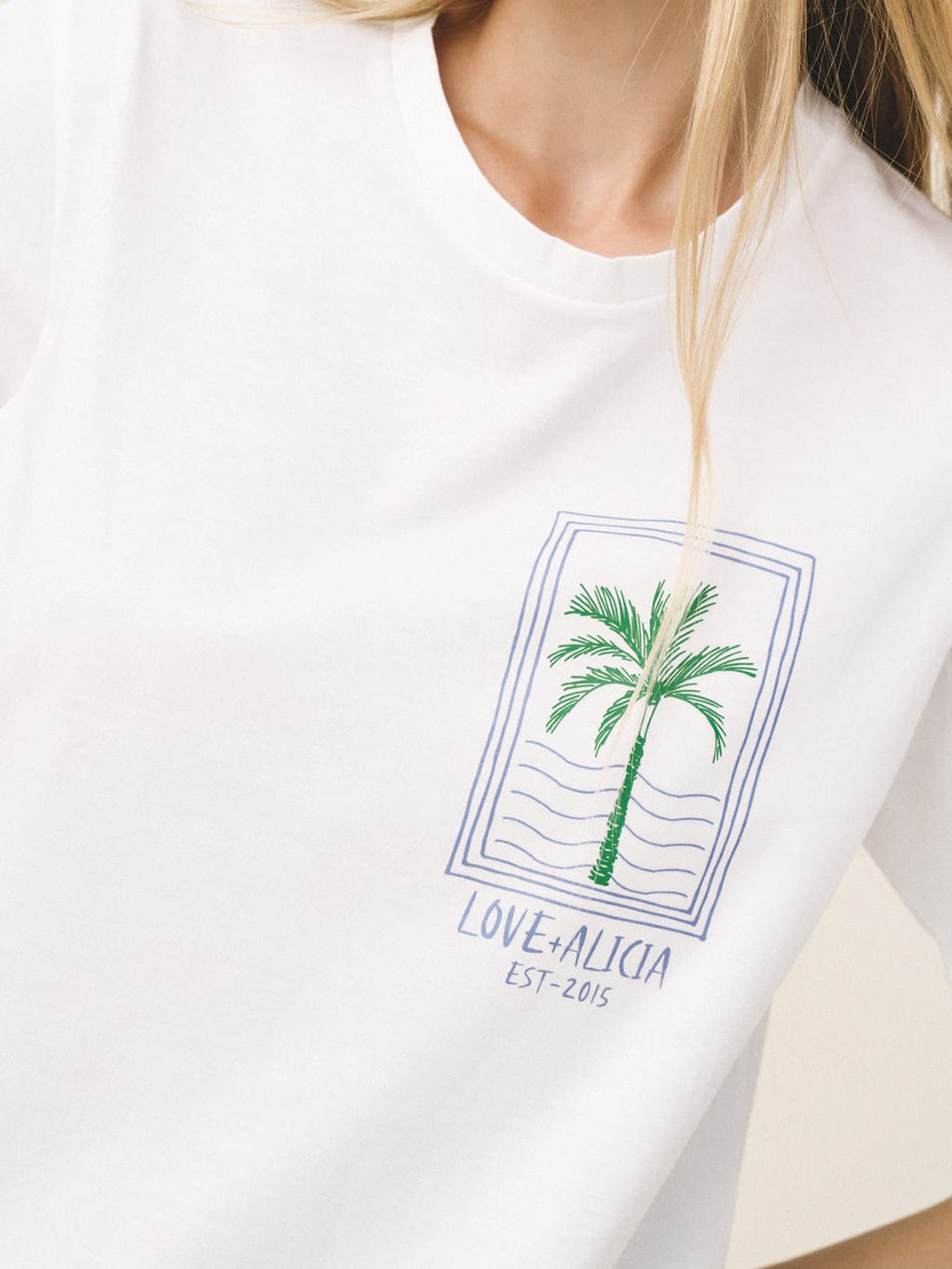 A close-up of a white crewneck t-shirt worn by a model, featuring a green palm tree graphic inside a blue rectangular border with the text 'LOVE+ALICIA EST-2016' printed below.