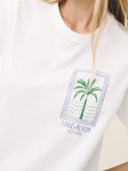 A close-up of a white crewneck t-shirt worn by a model, featuring a green palm tree graphic inside a blue rectangular border with the text 'LOVE+ALICIA EST-2016' printed below.