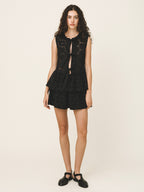 A model wearing a black sleeveless lace-trimmed top with keyhole cutouts and matching black shorts, paired with black Mary Jane shoes, against a plain light beige studio background.