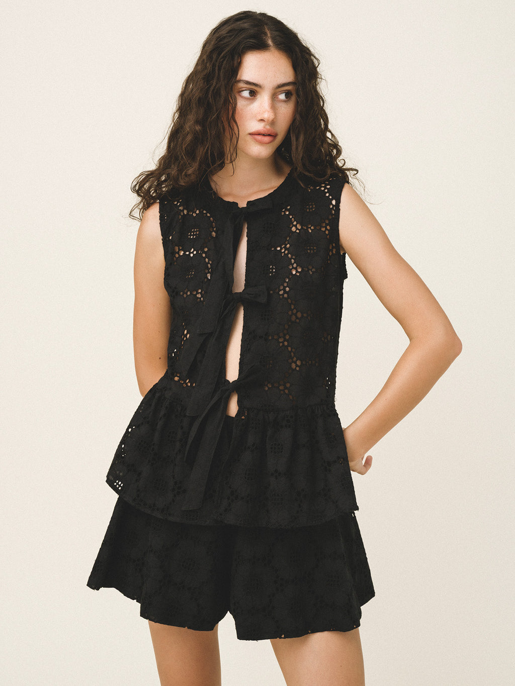 A woman modeling a black sleeveless lace top with a tiered ruffle hem and matching black shorts against a plain light beige studio background.