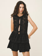 A woman modeling a black sleeveless lace top with a tiered ruffle hem and matching black shorts against a plain light beige studio background.