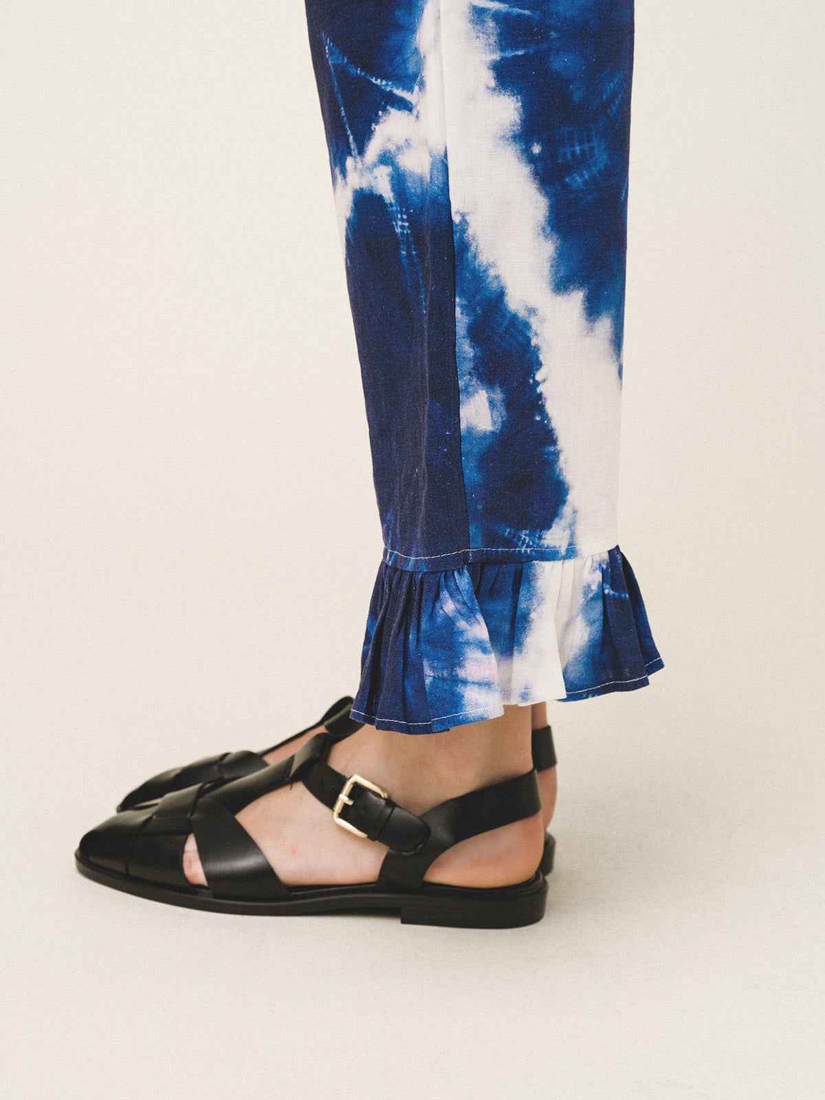 Blue tie-dye pants with ruffled hem worn by a person, paired with black sandals on a light background.
