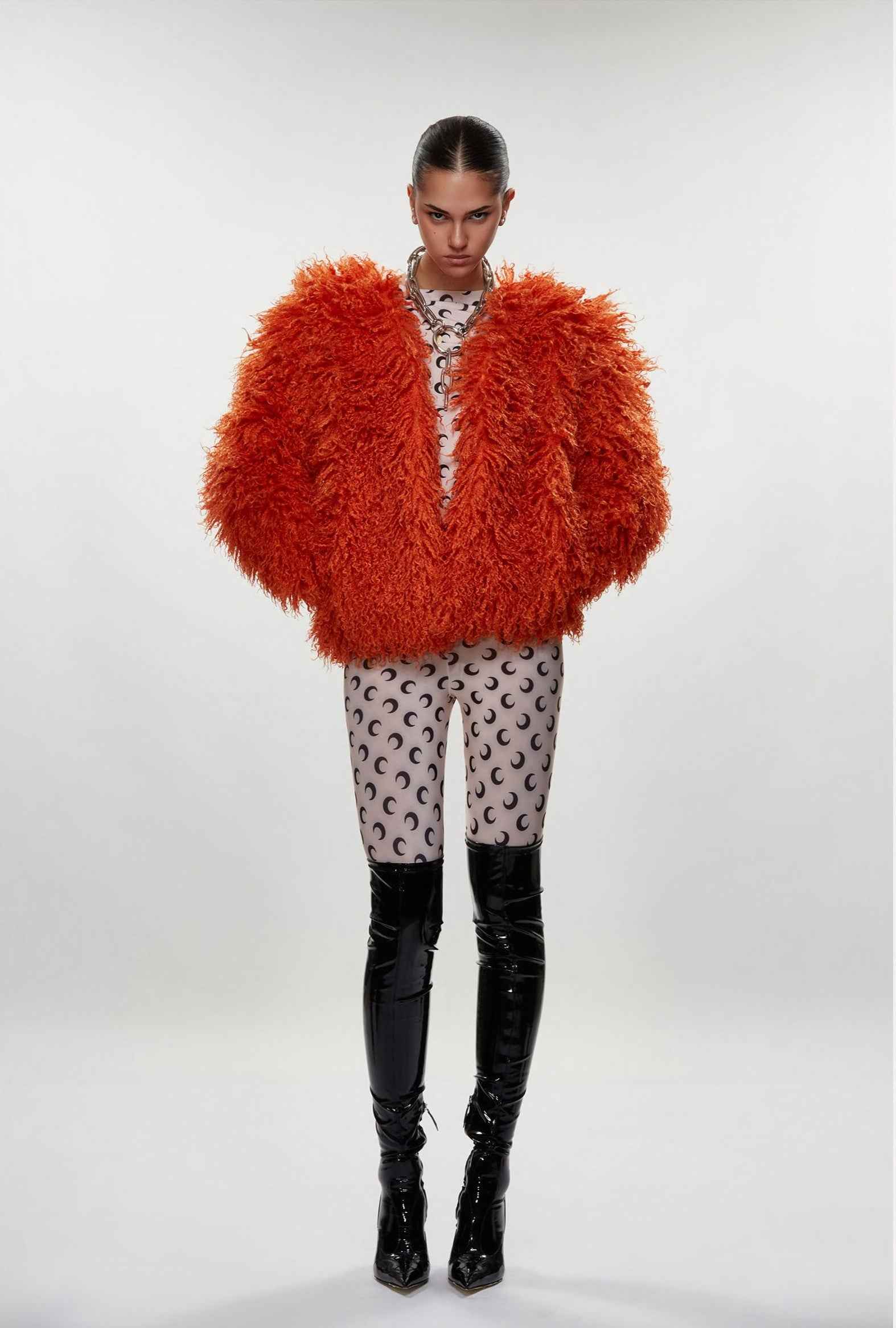 Person wearing a bright orange fluffy jacket with patterned pants and black boots on a white background