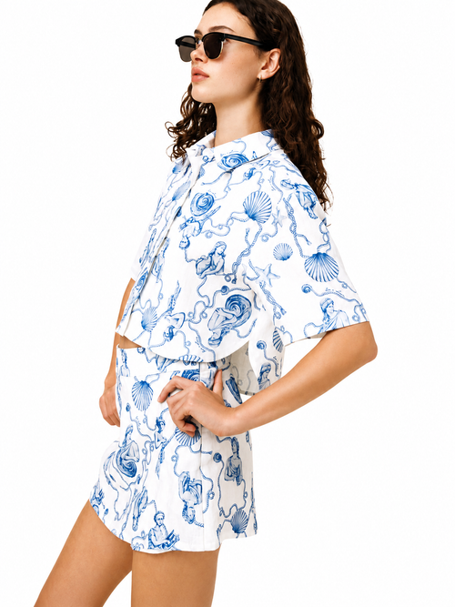 A woman modeling a white two-piece set with a blue seashell and sea creature print, featuring a short-sleeve crop top and matching shorts, set against a plain white background.