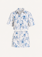 A matching two-piece set featuring a short-sleeve button-up shirt and a high-waisted mini skirt, both in white fabric with a blue toile-style print of sea creatures, shells, and waves. The set is displayed on a clean, solid off-white background.