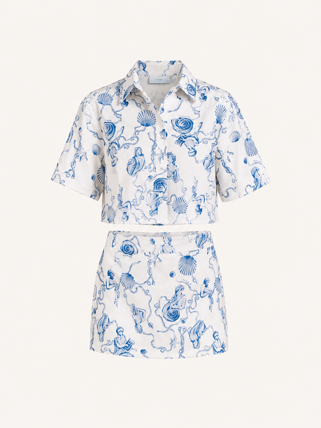A matching two-piece set featuring a short-sleeve button-up shirt and a high-waisted mini skirt, both in white fabric with a blue toile-style print of sea creatures, shells, and waves. The set is displayed on a clean, solid off-white background.