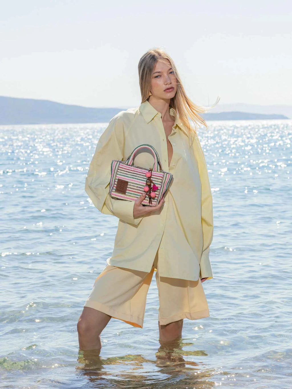 Woman in a light yellow outfit holding a striped handbag on a beach
