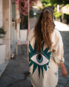 Person wearing a white "eye" see you jacket with a large eye design on the back, walking down a street.