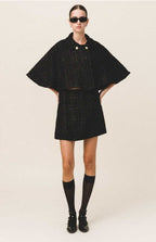 A model wearing a black tweed cape with gold button details and a flared silhouette, paired with a matching skirt.