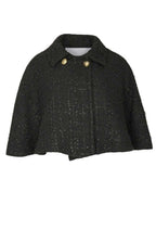 A model wearing a black tweed cape with gold button details and a flared silhouette, paired with a matching skirt.