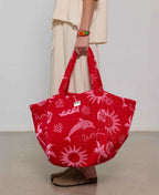 Red tote bag with pink designs held by a person wearing beige pants and brown shoes.
