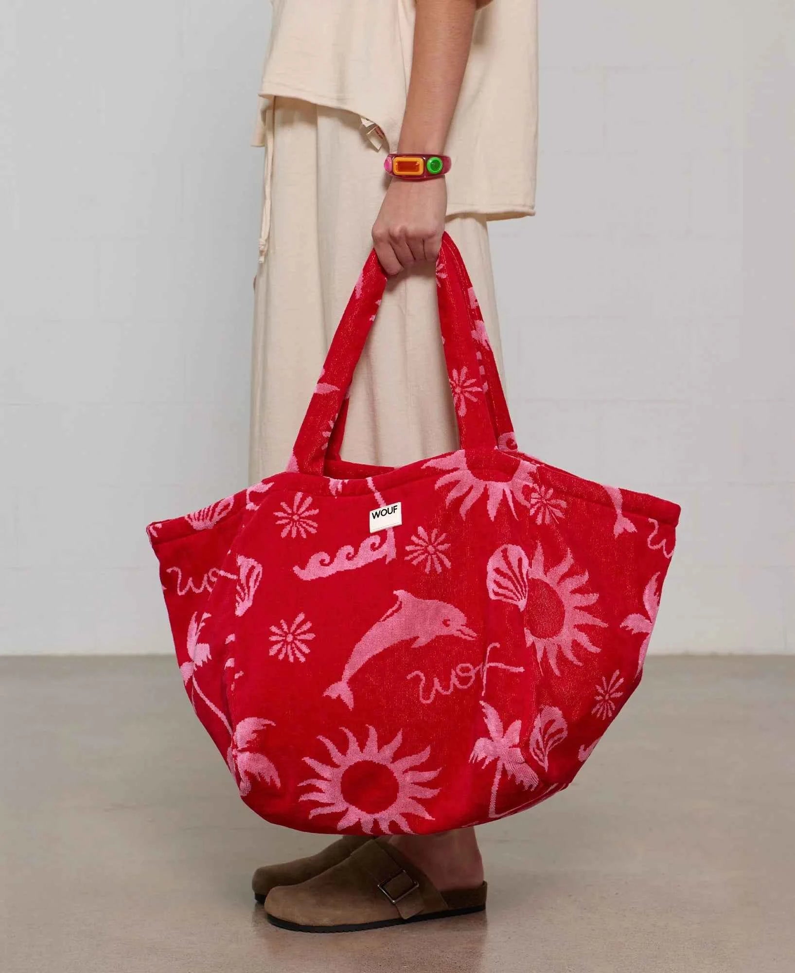 Red tote bag with pink designs held by a person wearing beige pants and brown shoes.
