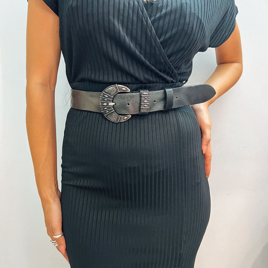 Person wearing a black ribbed dress with a belt featuring a large decorative buckle on a plain background