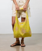 Person holding a yellow tote bag with a pattern, wearing beige shorts and black sandals on a plain background