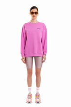 A model stands against a white background wearing a bright pink crewneck sweatshirt with a small embroidered logo on the left chest, matching pink biker shorts, white socks, and colorful sneakers.