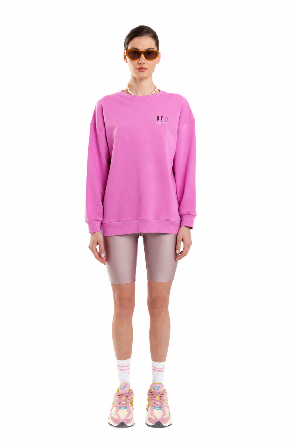 A model stands against a white background wearing a bright pink crewneck sweatshirt with a small embroidered logo on the left chest, matching pink biker shorts, white socks, and colorful sneakers.