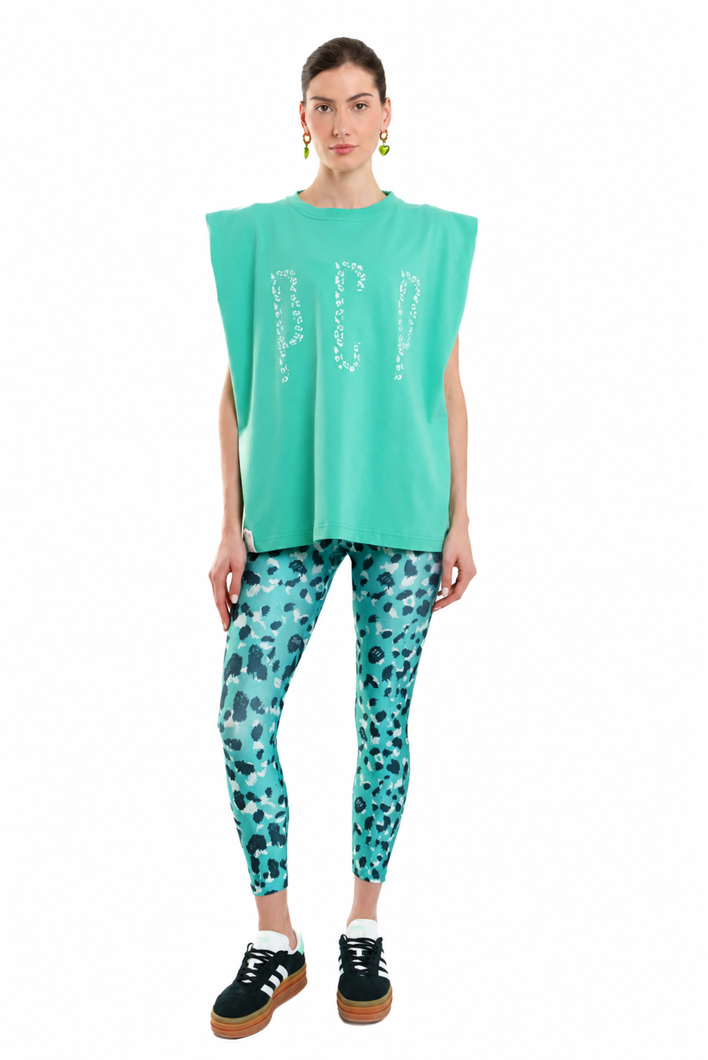 A model stands against a white background wearing a sleeveless mint green top with rhinestone lettering, matching patterned leggings, and black sneakers.