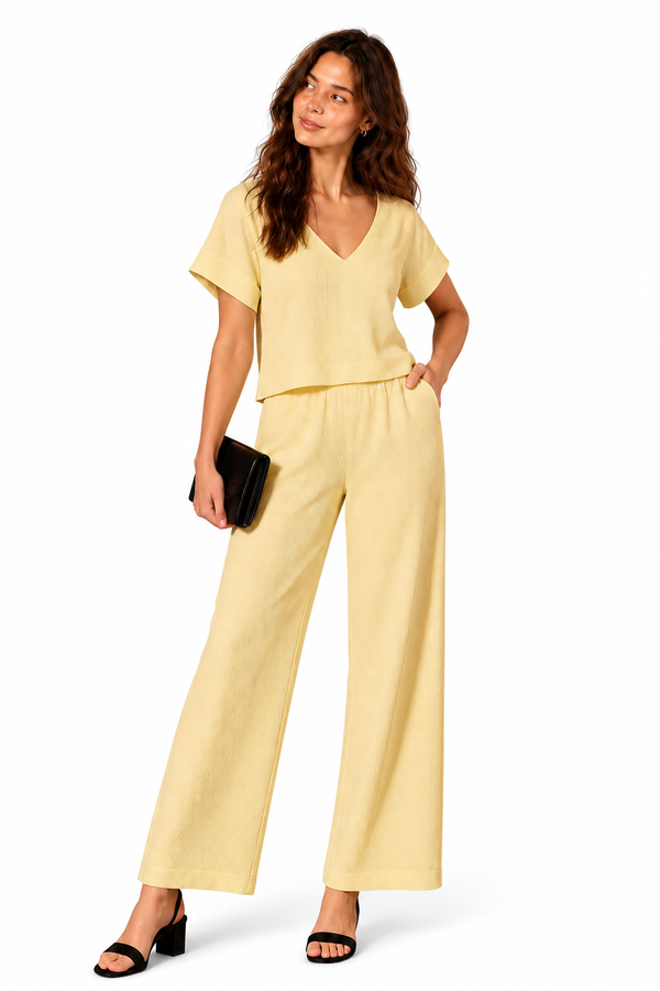 A woman modeling a pale yellow two-piece set consisting of a short-sleeve V-neck top and matching wide-leg pants, paired with black sandals and a small black clutch, on a white studio background.