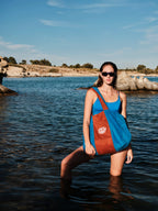 Woman standing in water wearing a blue tank top and carrying a red and blue bag.