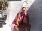 Woman sitting outdoors with a pink towel draped over her shoulders, surrounded by potted plants.