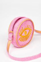 A pink textured crossbody bag with a yellow embroidered eye and the words 'SUN OF A BEACH' on the front, featuring tan leather trim and a matching strap, set against a plain white background.
