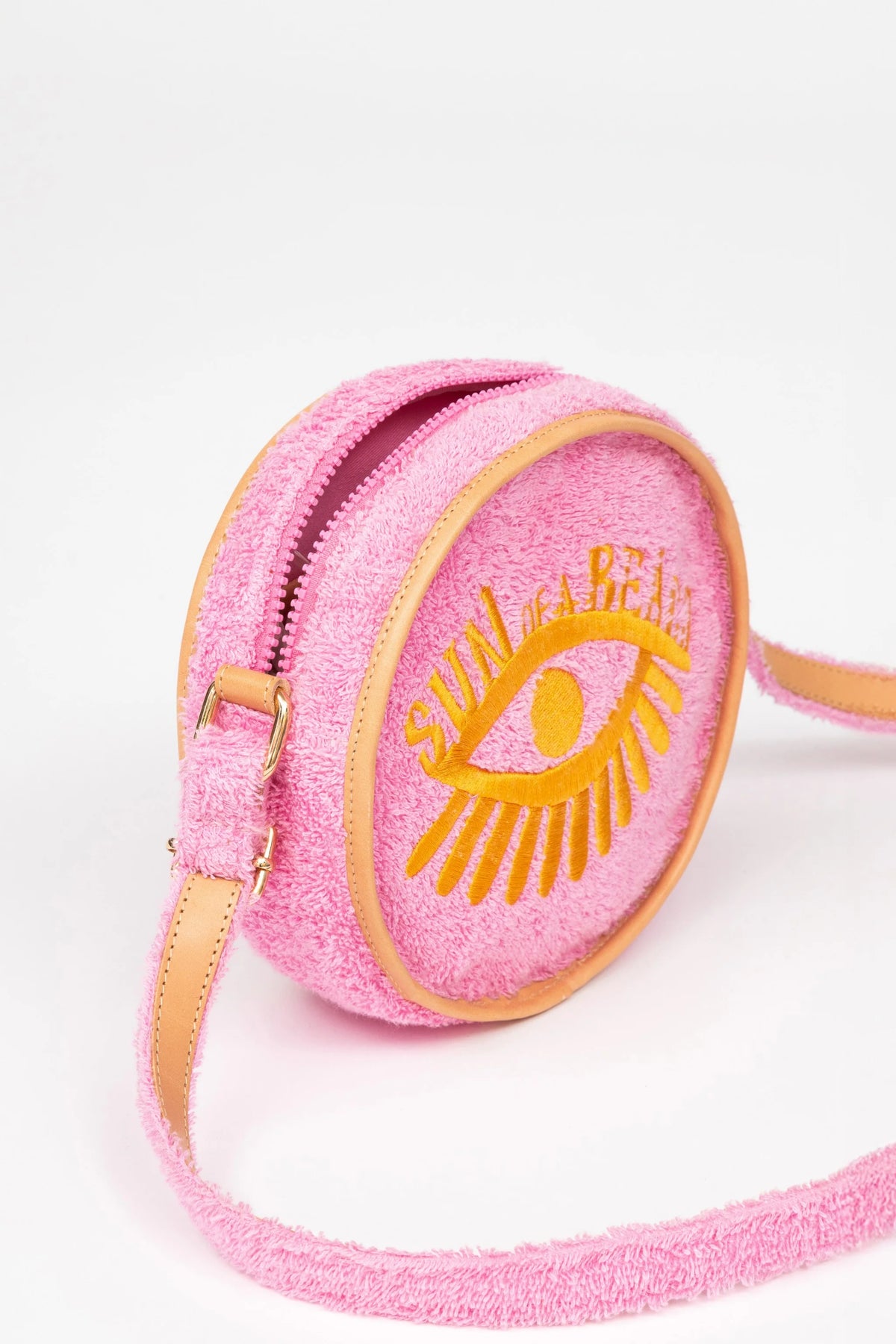 A pink textured crossbody bag with a yellow embroidered eye and the words 'SUN OF A BEACH' on the front, featuring tan leather trim and a matching strap, set against a plain white background.