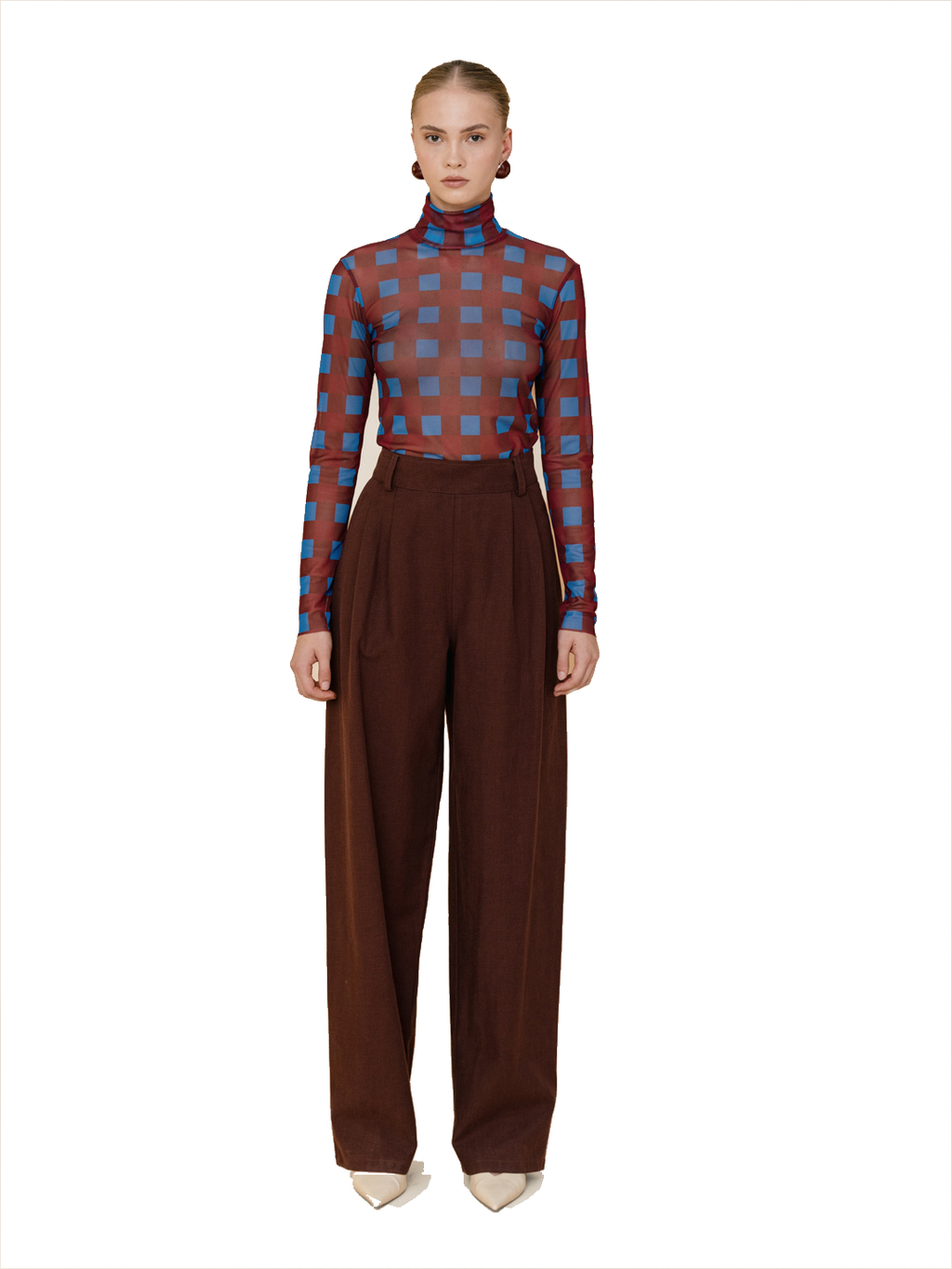A model stands against a white background wearing a sheer long-sleeve turtleneck top with a blue and brown checkered pattern, paired with high-waisted brown wide-leg trousers and beige pointed shoes.