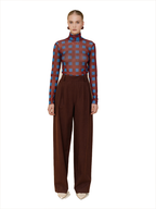 A model stands against a white background wearing a sheer long-sleeve turtleneck top with a blue and brown checkered pattern, paired with high-waisted brown wide-leg trousers and beige pointed shoes.
