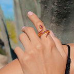 Hand wearing a gold ring with red gemstones against a blurred outdoor background