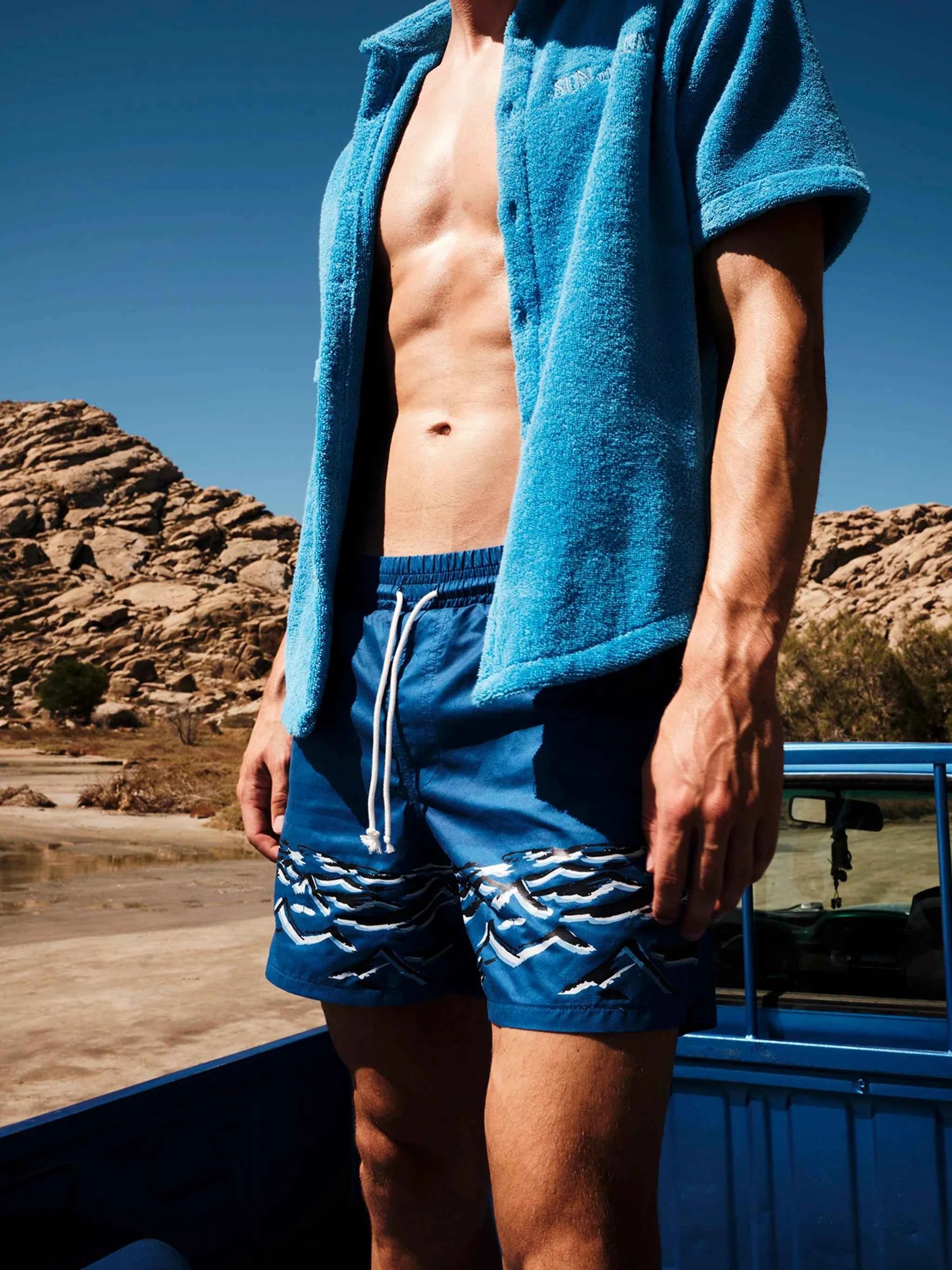 Person wearing a blue towel and swim shorts standing by a vehicle with a desert landscape in the background
